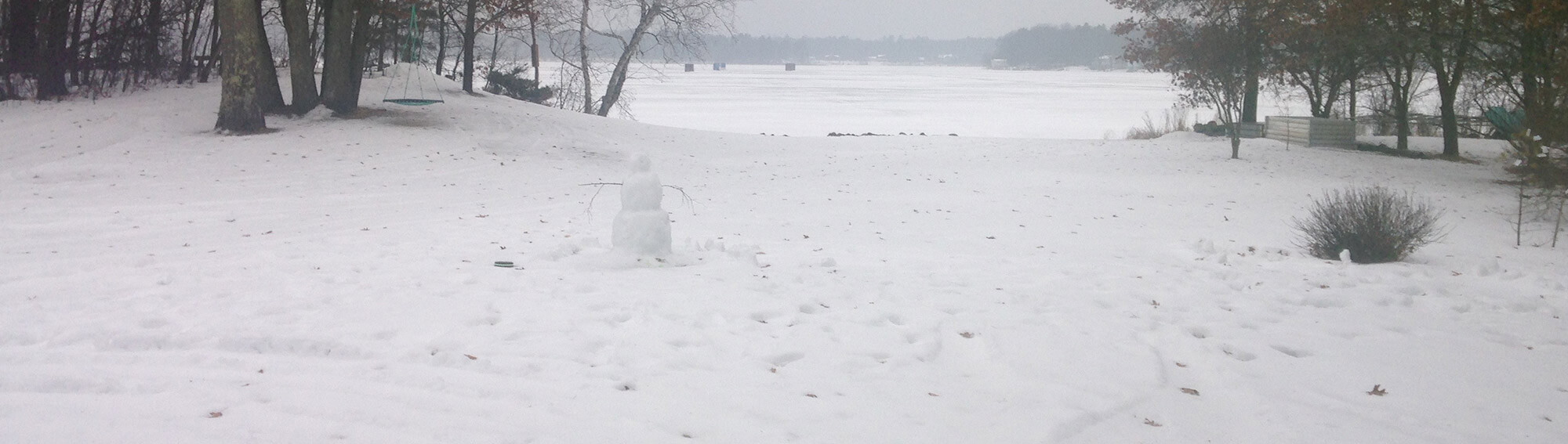 Photo of a winter landscape with a snowman in the foreground and ice fishing houses on a lake in the background